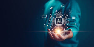 The image shows a person holding out their hand, with a glowing digital AI chip or processor graphic hovering above it. The AI chip has circuit-like connections spreading outward and is surrounded by various technology icons including cloud storage, settings, devices, and servers. The background has a teal/blue tone. Credit: Shutterstock